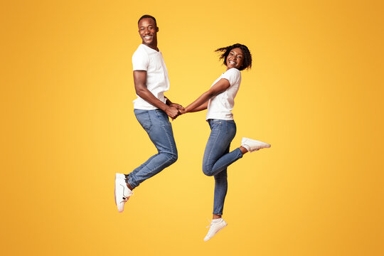 Emotional black couple in love holding hands and jumping up over orange studio background. Cheerful african american lovers young man and woman dancing, expressing love, happiness, joy