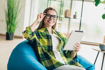Attractive young woman wearing glasses and using a tablet while relaxing in a cozy indoor space...