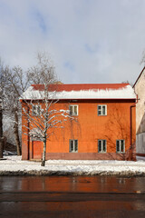 Orange residential building with snow-covered roof and bare tree in winter. Wet asphalt road and melting snow in urban environment