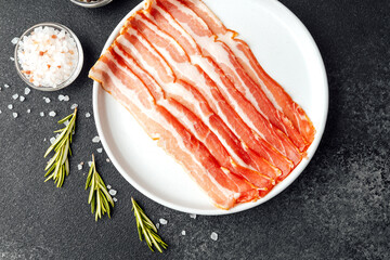 Raw bacon slices on white plate with pepper, salt and rosemary