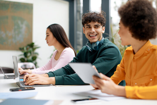 Happy diverse students sitting in classroom and communicating, using gadgets while being absorbed in studying, chatting and smiling, free space