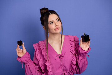 Elegant young woman wearing a pink ruffled dress holding perfume bottles against a purple...