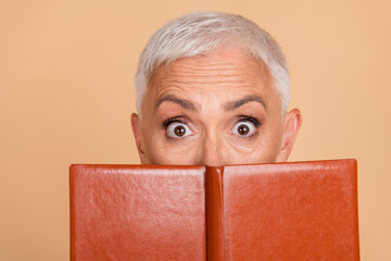 Charming mature woman holding a book in front of her face while expressing curiosity and excitement...