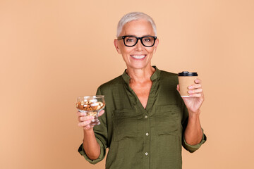 Smiling mature woman holding a coffee cup and a bowl of nuts, standing against a beige background,...