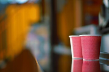 Two red paper cups placed on a reflective glass counter at a local beverage shop, creating a cozy and vibrant ambiance with a warm, blurred background with shallow depth of field