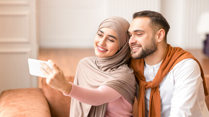 Young Arab Couple Making Selfie On Phone Sitting Together On Couch Having Fun At Home. Modern...