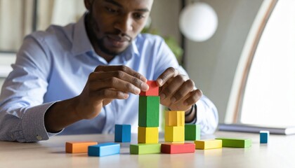 Fototapeta premium A man in a blue shirt carefully stacks colorful wooden blocks on a table, focusing on building a balanced structure.
