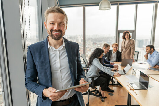 A businessman in a blue suit stands in a modern office, holding a tablet and smiling confidently. He looks directly at the camera. Several other people in business attire are sitting at a table