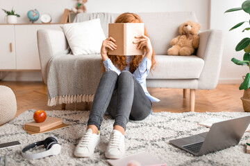 College Exams. Young red-haired teenage girl holding and covering face with paper book, hiding...
