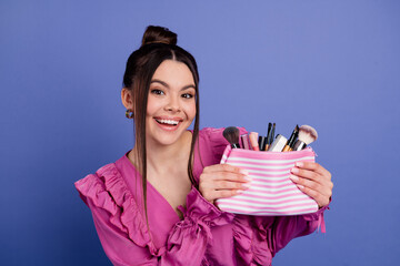 Smiling brunette woman holding a striped makeup bag with brushes against a purple background,...