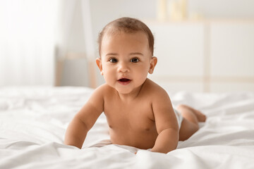 Portrait of cute small African American baby crawling on bed on the white blanket at home. Naked black infant lying on belly, looking at camera. Free copy space, selective focus, blurred background