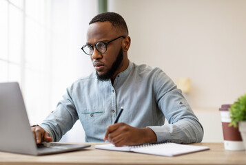 Fototapeta premium Serious Black Man Writing Business Report At Laptop Or Filling Tax Form At Workplace Sitting In Modern Office. Paperwork, Entrepreneurship Occupation And Career Concept