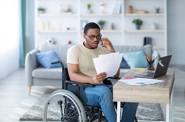 Online work for disabled people. Young black guy with documents speaking on phone in front of laptop at home. Millennial handicapped man in wheelchair having remote job, communicating on mobile device