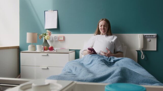 Full shot of cheerful young Caucasian woman in white medical gown sitting in bed in maternity hospital ward, typing messages on smartphone, then having labor contractions and grimacing in sharp pain