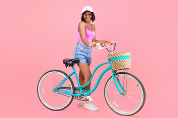 Full length of happy black lady in summer outfit standing with classic bicycle on pink studio...