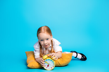 Smiling red-haired girl holding a clock while lying on a pillow, set against a bright blue background, showcasing childhood charm