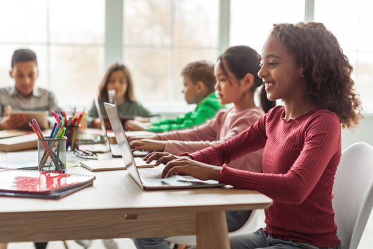 Online Learning. Black Schoolgirl Using Laptop Sitting At Desk With Multiethnic Classmates In Classroom Indoor. Modern School Education And Technology Concept. Selective Focus
