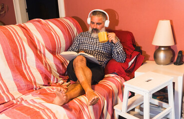 Fototapeta premium Relaxed senior man reading a book, drinking tea and listening to music on sofa