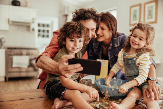 Happy young family taking a selfie with a smartphone while being messy and baking with flour at home in the kitchen