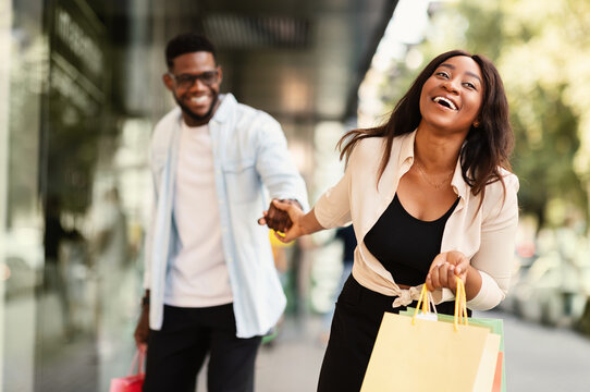 Sales Season. Portrait Of Smiling Excited African American Lady Pulling Boyfriend To Shopping Store To Buy New Clothes, Fooling And Running To Mall Together. Discount, Fun, Purchase And Retail - Powered by Adobe