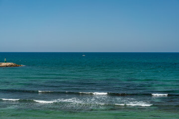 Blue Mediterranean sea with gentle waves and a green lighthouse on the breakwater. Summer coastal scene with a distant sailing boat and layered water texture.