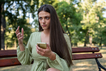 Confused young woman using smartphone while sitting on park bench, thoughtful expression in natural setting