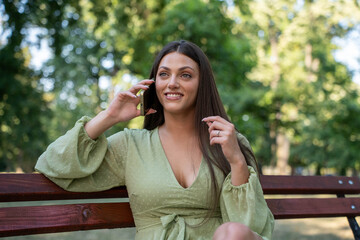 Beautiful young woman talking on smartphone in green park, smiling and enjoying relaxing morning outdoor atmosphere