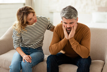 Middle Aged Wife Comforting And Supporting Depressed Husband Sitting On Couch At Home. Couple...
