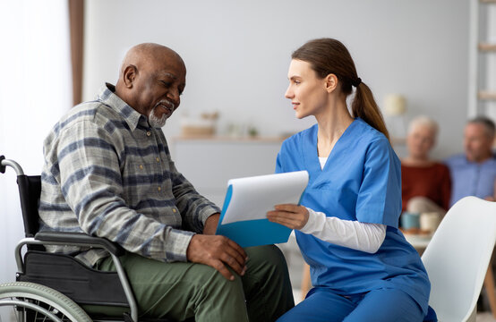 Pretty young woman in workwear nurse helping black senior man in weheelchair filling papers, cheerful elderly african american male patient signing documents at nursing home