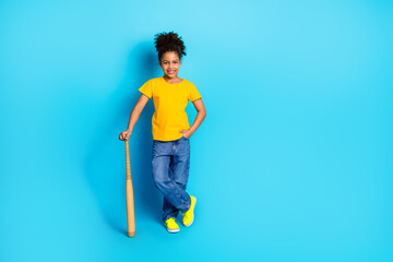 Portrait of a smiling young girl dressed casually in yellow and blue, standing against a vibrant blue background with a bat.