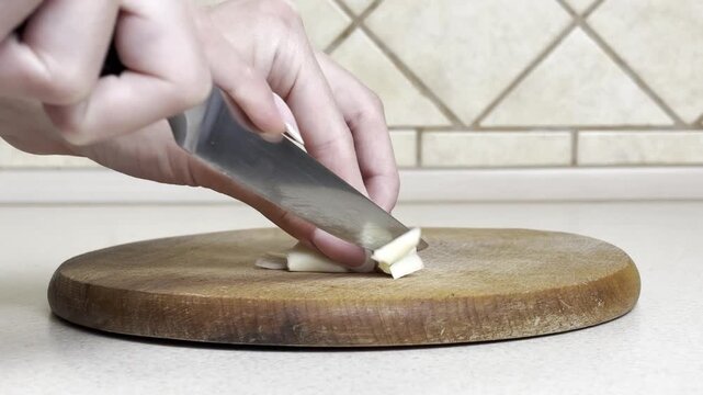 Close-up of woman's hand finely chopping garlic with a kitchen knife on wooden board, cooking preparation, fresh ingredients, healthy food, home kitchen, natural light, culinary process