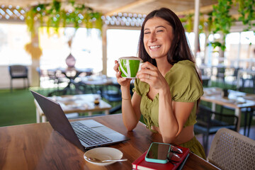 Professional woman sipping cappuccino while typing on laptop, embracing digital workspace in modern cafe setting