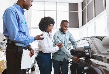 Car sales business. Manager talking to Afro couple, showing them new auto at dealership shop. Young...