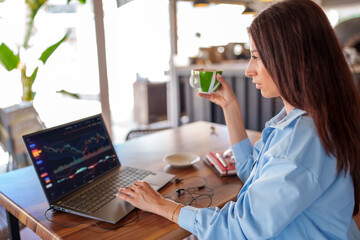 Businesswoman drinking coffee and analyzing stock market data on laptop, working remotely in cafe