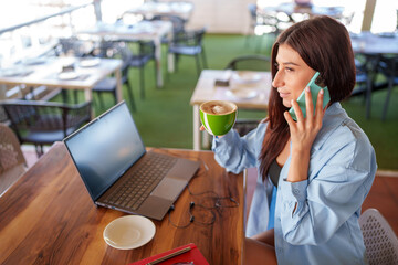 Young manager drinking cappuccino and talking on smartphone while telecommuting on laptop in cafe