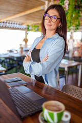 Professional woman crossing arms near laptop, drinking coffee while working remotely at restaurant table
