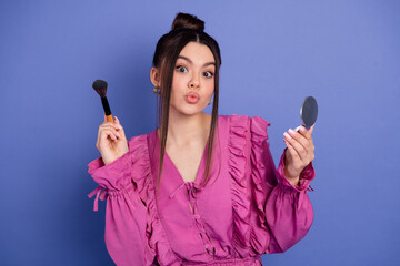 Beautiful young woman in a pink ruffled dress holding a makeup brush and mirror against a purple...