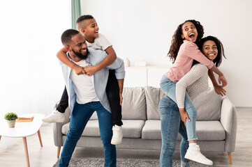Positive black mom and dad having fun with their kids, playing in living room, giving smiling son and daughter piggyback ride. African American family of four people enjoying spending time together