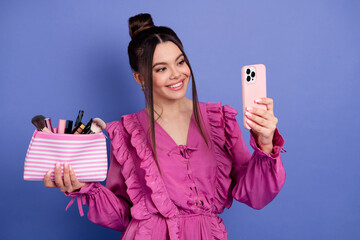 Smiling young woman in fashionable pink dress takes a selfie while holding makeup essentials...
