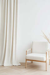 A wooden chair with white cushions is placed next to a long curtain and dried grasses on a light wooden floor. Minimalist interior design