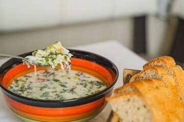 A vibrant bowl of cold okroshka soup filled with herbs and vegetables is placed on a dining table...