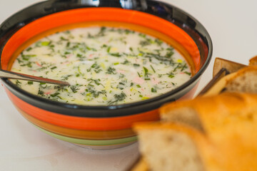 A vibrant bowl of cold okroshka soup filled with herbs and vegetables is placed on a dining table...