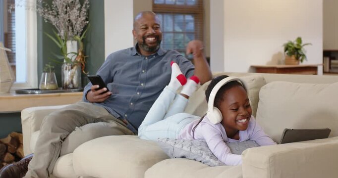 African American father and daughter hearing giggle, tickling feet and sharing tablet on sofa