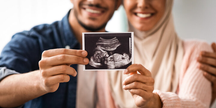 Closeup shot of happy pregnant muslim couple demonstrating baby sonography photo while relaxing together at home, showing first photo of their child, enjoying future parenthood, cropped image