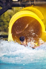 A young person is seen exiting a yellow water slide into a pool, creating a splash of water as they...