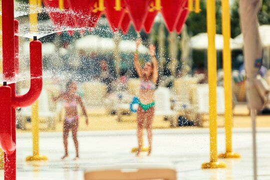 Two girls in swimsuits stand at a splash pad in a water park as water droplets from a bucket tower fall all around, creating a blurred, playful summer memory
