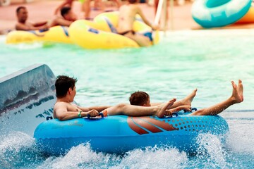 Two boys are riding on a blue inflatable raft down a lazy river. The water splashes around them as...