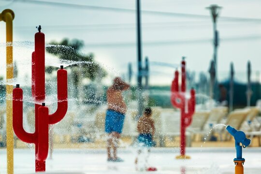 Amidst vibrant cactus-shaped sprinklers, a father and child delight in a refreshing splash pad under a hazy afternoon sun, creating a playful summer experience at the water park