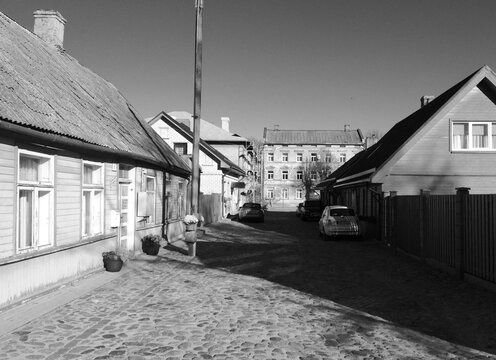 Fototapeta Black and white photo of a narrow cobblestone street with old wooden houses, parked cars, and a distant brick building under a clear sky.