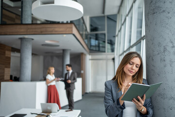 Businesswoman writing notes in modern office lobby with colleagues discussing in background